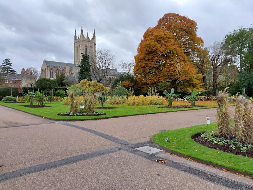 A well-maintained public garden with a neatly mowed lawn and a paved pathway weaving through the landscaped beds. The garden features a variety of plants, including ornamental grasses, small shrubs, a