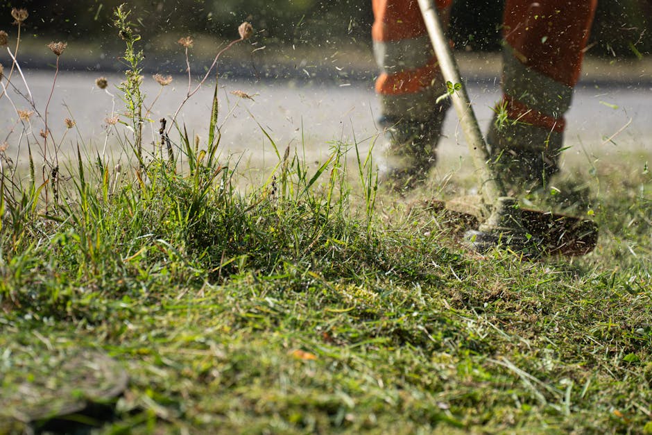 Close-up view of a gardener's legs and feet wearing orange work trousers and boots, operating a garden tiller on a patch of soil with patches of grass and weeds. The soil appears moist and freshly tur