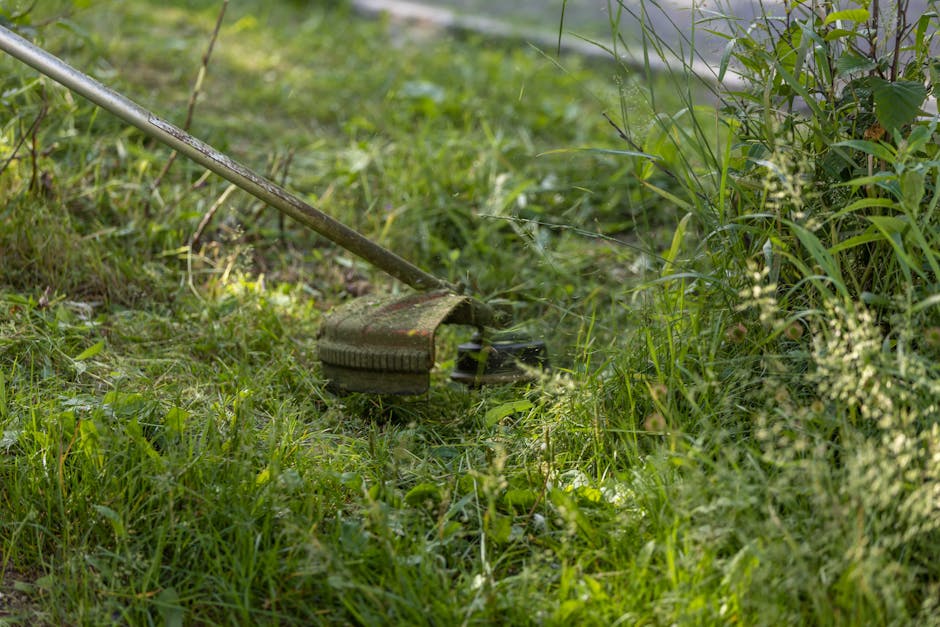 Close-up view of a gardener's legs and feet wearing orange work trousers and boots, operating a garden tiller on a patch of soil with patches of grass and weeds. The soil appears moist and freshly tur